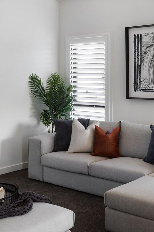 A bright, contemporary living room featuring a large light-grey sectional sofa with neutral cushions and a rust-coloured accent cushion. White plantation shutters, pale walls, dark carpet, a potted palm, and monochrome framed artwork create a clean, refined interior.