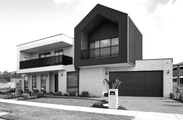 Black-and-white photograph of a contemporary multi-storey suburban home. The facade combines pale brickwork, smooth rendered surfaces, extensive dark-framed glazing, a projecting dark box-like upper level with a deep geometric opening, vertical fins, balconies, and an integrated double garage. Low-maintenance front landscaping and a paved driveway are visible.