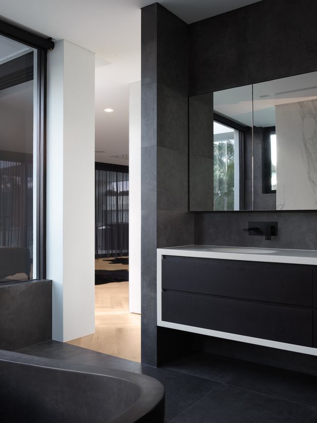 A sophisticated contemporary bathroom featuring a dark freestanding bathtub in the foreground, dark stone-look wall and floor finishes, a floating black vanity with a white benchtop, mirrored cabinet doors, and matte-black tapware. A bedroom or living area is visible beyond the open doorway.