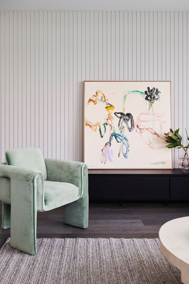 A refined contemporary living room featuring a pale green upholstered armchair, dark low-profile media or storage cabinetry, grey textured flooring, a neutral rug, and a large abstract artwork. The main feature wall is clad in narrow vertical fluted panels, creating a softly textured backdrop. A round light stone or pale surface is partially visible in the foreground, with a small plant and decorative vase adding natural detail.