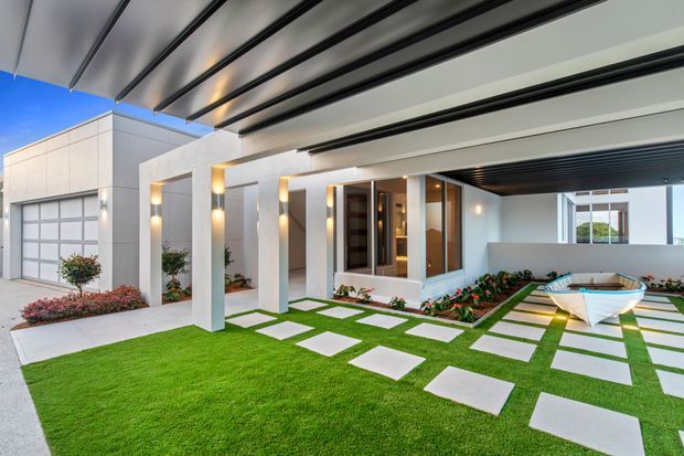 Contemporary residential front courtyard and covered entry viewed from outdoors. The home features a crisp white rendered facade, large garage door, glazed entry volume, deep covered canopy with dark exposed beams, geometric stepping-stone paths set into manicured lawn, and landscaped garden beds.