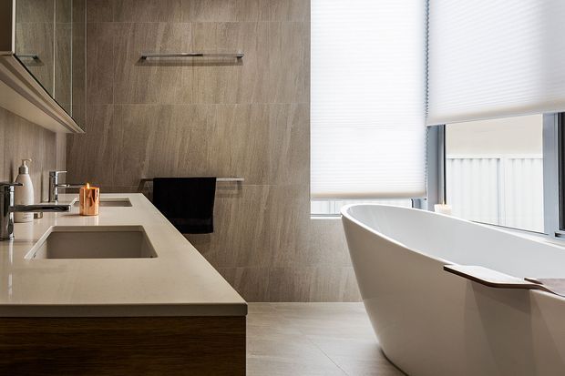 Contemporary bathroom featuring a long floating vanity with a pale stone benchtop, integrated sink, dark timber cabinetry, and warm grey wood-look wall tiling. A freestanding white bathtub sits beside large windows with translucent roller blinds, creating a bright, refined space.