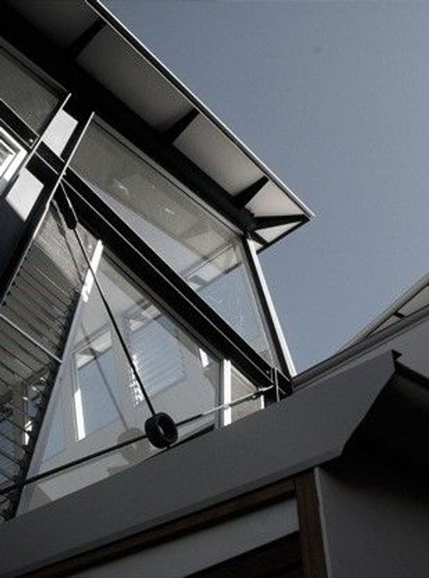 Upward-looking exterior architectural detail showing a modern multi-level glazed facade, dark structural framing, projecting roof eaves, and a glass balustrade around an elevated balcony or void. The composition emphasizes sharp angular lines, extensive glazing, and exposed structural elements against the sky.