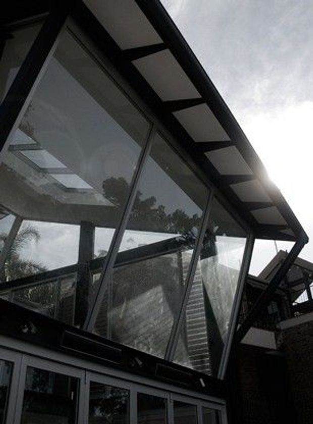 Upward-looking exterior view of a contemporary glazed architectural structure with steeply angled glass panels, dark structural framing, and a projecting roof canopy. Reflections show surrounding trees and adjacent building elements, while sunlight enters from the upper right.