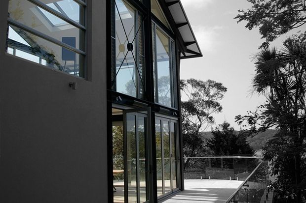 Exterior view of a contemporary multi-level home with extensive floor-to-ceiling glazing, dark metal window and door frames, a projecting roof eave, and a broad paved terrace or balcony overlooking a wooded landscape. The image also reveals parts of the bright interior through the glazing.