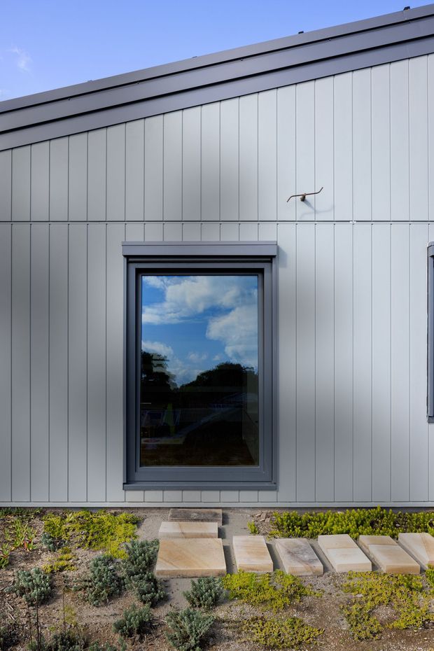 Exterior view of a contemporary single-storey building clad in pale grey vertical panels, with dark grey metal roof trim and a dark-framed window reflecting blue sky and trees. A paved stepping-stone path crosses a low, drought-tolerant landscaped garden in the foreground.