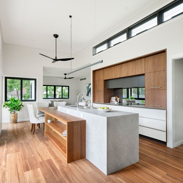 Bright open-plan kitchen and dining area with a large island, white waterfall benchtop, warm timber cabinetry and flooring, black-framed windows, and high clerestory glazing. The room includes an integrated dining table, contemporary pendant and ceiling fans, with greenery visible outside.
