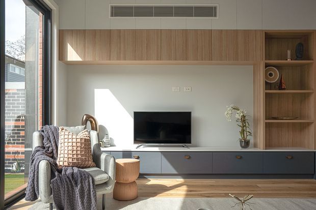 Bright contemporary living room with a built-in timber entertainment wall and shelving, dark blue-grey low cabinetry, television, armchair, woven ottomans, and large glazing opening toward an outdoor area. Natural light creates a calm, warm, refined interior.