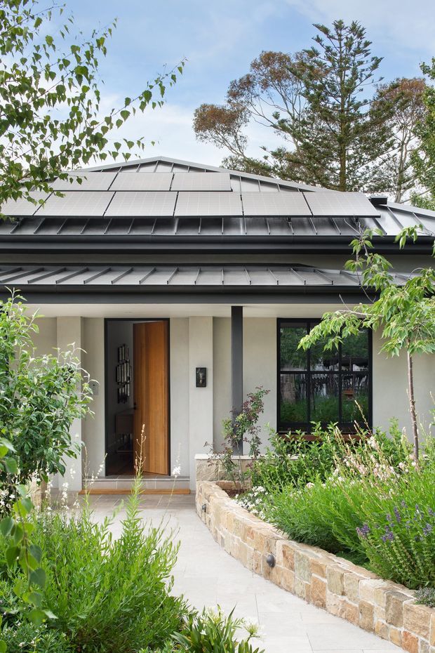 Front exterior view of a contemporary single-storey home, showing a layered dark metal roof with prominent solar panels, pale rendered walls, a tall timber entry door, black-framed glazing, and a landscaped garden with a low natural stone retaining wall and paved path.