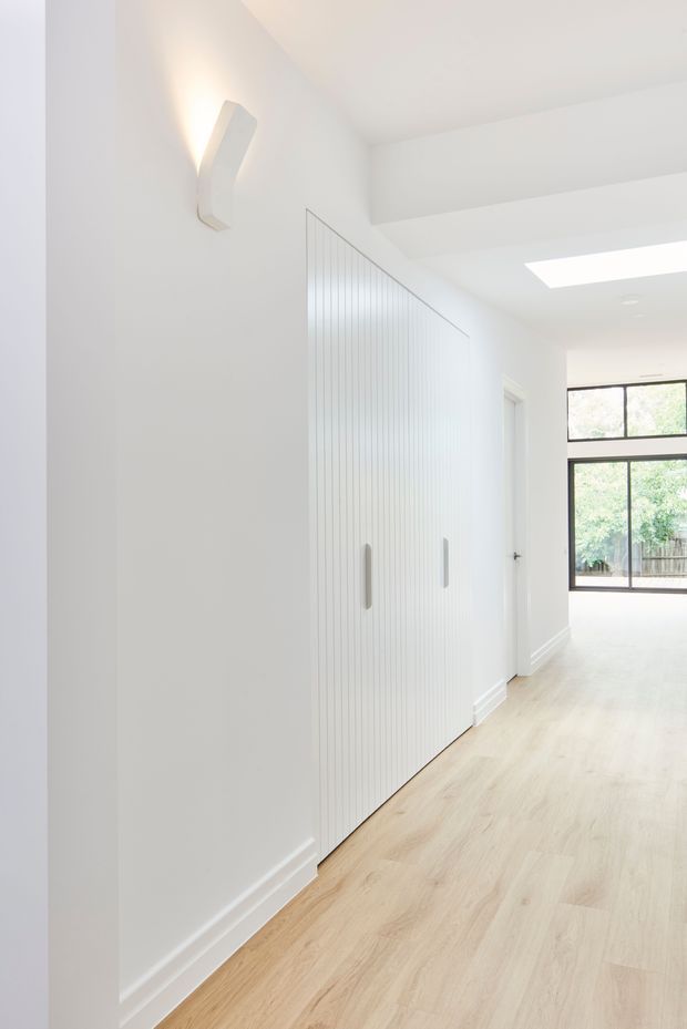 Bright minimalist hallway with white walls and ceiling, pale timber flooring, a large fluted built-in storage cupboard with vertical panel detailing and simple handles, a wall-mounted sculptural light, and black-framed glazing at the far end. A skylight or rooflight adds natural illumination.