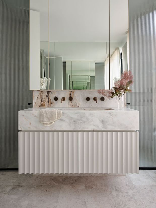 A refined contemporary bathroom centred on a sculptural double vanity. The vanity and integrated basin are clad in pale veined stone, with a fluted white cabinet base and dark bronze tapware. A large mirrored wall expands the space, while vertically ribbed translucent wall panels and pale stone flooring add texture. Soft floral arrangements provide a subtle decorative accent.