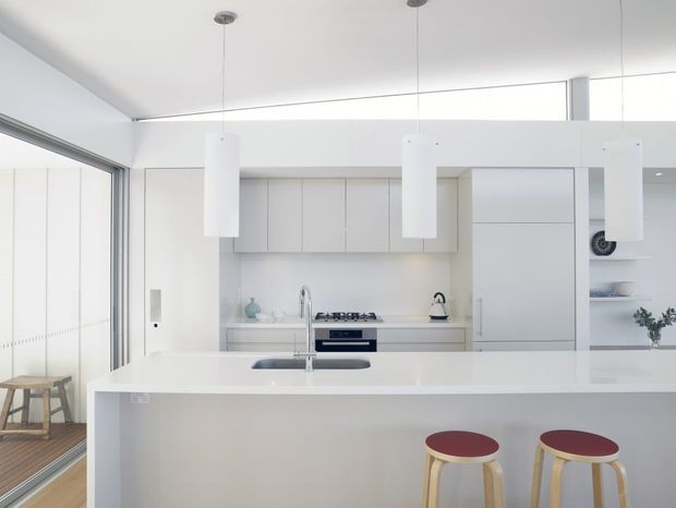 Bright minimalist contemporary kitchen with a large white island, integrated sink, pale cabinetry, white benchtops and three suspended cylindrical pendant lights. Two timber stools with burgundy seats sit at the island. Full-height glazed doors open toward a timber-floored outdoor area, while open shelving and a concealed appliance wall complete the clean, streamlined fit-out.