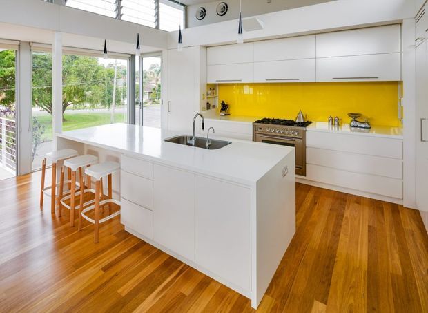 Bright contemporary kitchen with a large white island incorporating a stainless-steel sink and seating for four, white handleless cabinetry, a vivid yellow glass splashback, stainless-steel range cooker, timber flooring, and extensive windows and glazed doors opening toward a green outdoor setting. Slim pendant lights and high-level louvred glazing add architectural detail.