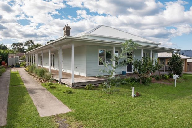 Single-storey light-coloured weatherboard house with a broad hipped metal roof, covered verandah and simple white posts. The home sits on a flat suburban garden with a concrete path, lawn and modest planting; neighbouring homes are visible in the background under a partly cloudy sky.