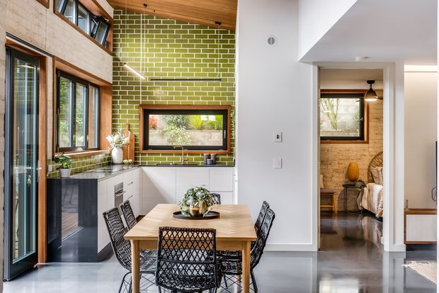 Bright open-plan dining area beside a compact kitchen, featuring a timber dining table with woven black chairs, green glazed brick splashback, white cabinetry, dark polished flooring, timber-framed glazing and a warm timber ceiling. An open doorway leads to a bedroom or living space with exposed brick walls.