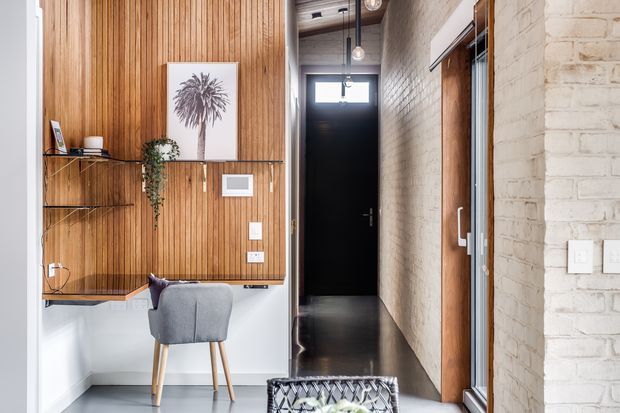 A narrow, stylish residential entry hall with a compact desk or console nook. The space features a full-height vertical timber slat feature wall with floating shelves, a small upholstered chair, framed botanical artwork, and a trailing plant. White-painted brick walls, dark polished flooring, black interior doors, exposed pendant lighting, and warm timber joinery create a refined industrial-contemporary look.