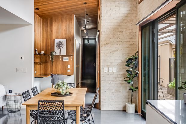 Bright contemporary dining area with a light timber dining table and woven black chairs, adjacent to a white-painted exposed brick wall and warm timber wall panelling. The room features a timber-lined ceiling, pendant lighting, indoor plants, open shelving, and large black-framed glazed doors opening to an outdoor area.