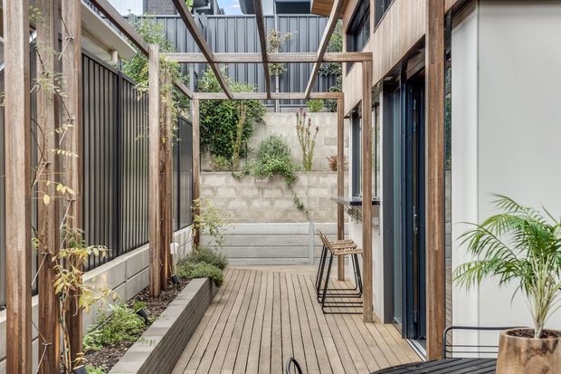 A narrow, landscaped outdoor courtyard or side passage with a timber deck, raised planter beds, climbing greenery, timber pergola framing, and contemporary glazed openings along the right side. A concrete block boundary wall and built-in bench complete the secluded garden space.