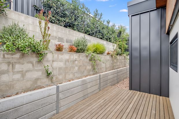 A narrow landscaped outdoor passage beside a modern building, featuring a timber deck, stepped concrete block retaining walls with integrated planting beds, and a dark vertical-panel structure with timber soffit detailing. The space is bright and contemporary, with layered greenery along the boundary.