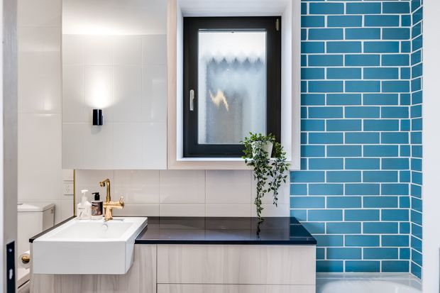 Bright contemporary bathroom featuring a floating white vanity with integrated basin, pale timber-look cabinetry, a dark benchtop, glossy white wall tiles, and a striking turquoise-blue brick-pattern tiled feature wall around the frosted window and bathtub. Black window framing and a small trailing plant add contrast.