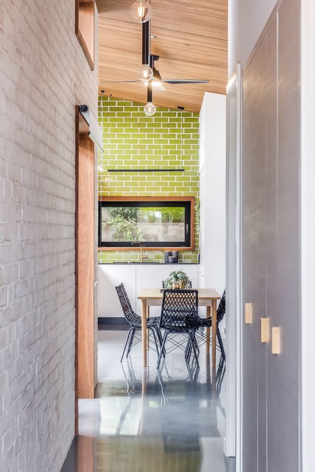 A narrow contemporary dining and kitchen area viewed from a hallway, featuring a vivid lime-green glazed brick feature wall, white cabinetry, a timber dining table with black woven chairs, polished concrete flooring, and a warm timber slatted ceiling. Exposed pale brickwork lines the hallway, while pendant lights and a ceiling fan add functional detail.