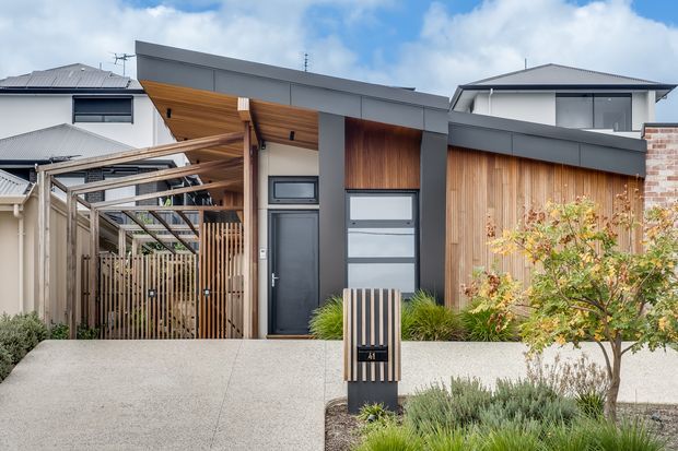 Contemporary residential facade viewed from the street, featuring a low-pitched metal roof, dark grey rendered wall planes, warm vertical timber cladding, a timber-screened covered entry, and a modern garage door. A landscaped front garden and concrete driveway complete the composition.