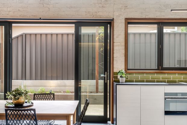 Bright contemporary dining and kitchen interior with exposed light-painted brick walls, dark-framed glazed sliding doors, and a compact white kitchen. The dining area includes a pale rectangular table and black woven chairs, while the kitchen has white flat-panel cabinetry, a dark benchtop, and a green subway-tile splashback. Windows and doors open toward a fenced outdoor courtyard.