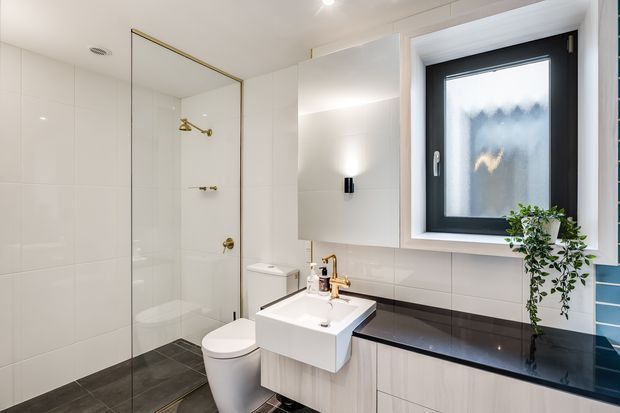 Bright contemporary bathroom with a frameless glass shower enclosure, white wall tiles, dark grey floor tiles, wall-mounted white vanity, black stone benchtop, brass tapware and shower fittings, and a black-framed frosted window with a trailing plant.