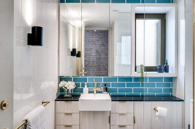 Bright contemporary bathroom with a wall-mounted white rectangular basin, pale timber vanity cabinetry, glossy deep teal subway-tile splashback, large mirrored cabinetry, white wall tiles, black wall sconces, and a dark benchtop. A window provides natural light, while a partial reflection reveals a textured brick wall beyond.