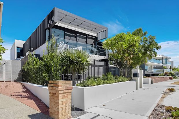 A contemporary multi-level residence with a strong geometric facade, dark vertical cladding, white rendered elements, extensive glazing, and a rooftop terrace or balcony with a pergola-like projecting canopy. The home is set behind landscaped front gardens and low rendered and brick boundary walls, with a paved driveway and footpath visible in the foreground under a clear blue sky.