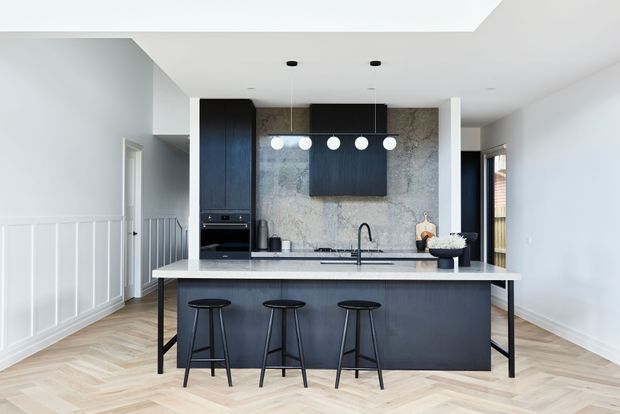 Bright contemporary kitchen viewed from the living or circulation area, featuring a large dark island with three stools, pale stone benchtops, a textured stone splashback, dark integrated cabinetry, pendant lighting, and light timber herringbone flooring. White wall panelling and a stair or hallway area are visible to the left.