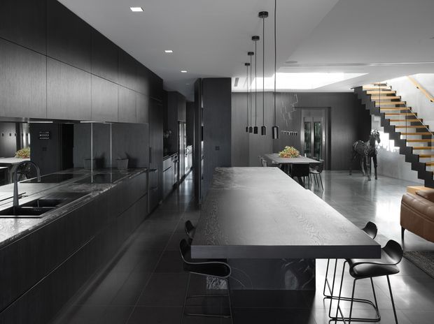 A dramatic open-plan contemporary kitchen and dining area with dark full-height cabinetry, a large stone island with integrated sink, black pendant lights, and bar stools. The space opens toward a dining table and a sculptural staircase with timber treads and dark stringers, creating a refined monochromatic interior.