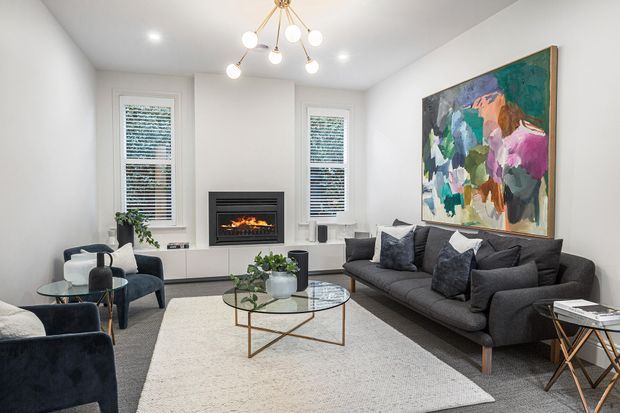 Bright, refined living room with a large dark sectional sofa, two lounge chairs, a glass coffee table with a brass-toned geometric base, a pale area rug, a contemporary fireplace, white walls and window shutters, and a bold abstract artwork. A sculptural multi-globe pendant provides a decorative focal point.