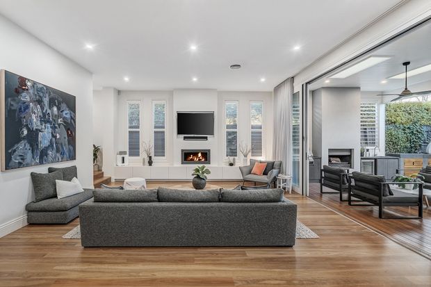 Bright contemporary living room with grey sofas, timber flooring, a wall-mounted television, fireplace, recessed ceiling lights and full-height sliding glass doors opening to an adjoining lounge or outdoor area. White plantation shutters and neutral furnishings create a clean, refined interior.