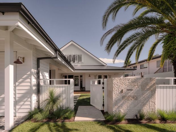 A bright coastal-style residential exterior with white weatherboard-clad buildings, pitched gabled roofs, covered entry areas, and a low white boundary fence. A textured rendered masonry wall forms the gate entry, with a paved path, lawn, ornamental grasses, and a large palm tree contributing to the landscaped frontage.