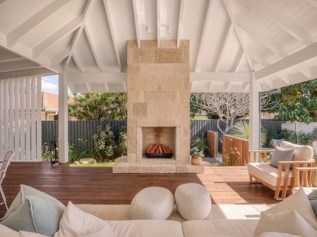 A bright, airy covered outdoor living area with a high white timber-framed vaulted ceiling and exposed rafters. A large stacked-stone fireplace forms the centrepiece, opening onto a timber deck with relaxed upholstered seating, a light rug, and sculptural ottomans. Vertical privacy screening, fencing, planting, and an outdoor dining or lounge zone are visible around the perimeter.