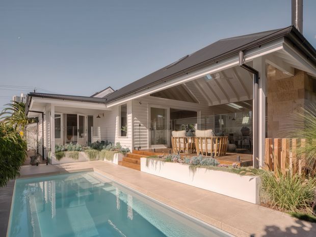Exterior view of a bright, single-storey residential home surrounding a narrow swimming pool. The house features light horizontal weatherboard cladding, a dark pitched roof, white trim, large glazed openings, and a covered outdoor dining area with exposed painted rafters. Landscaping and timber screening frame the pool terrace.