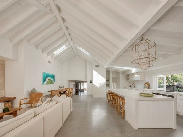 Bright open-plan interior combining a living room, dining area, and kitchen beneath a dramatic white pitched ceiling with exposed beams and rafters. The space features polished grey concrete flooring, white cabinetry and walls, a large kitchen island with timber stools, a cream sectional sofa, timber lounge furniture, skylights, and expansive glazing. A sculptural pendant light hangs above the kitchen, with a partial stone wall visible at the left.
