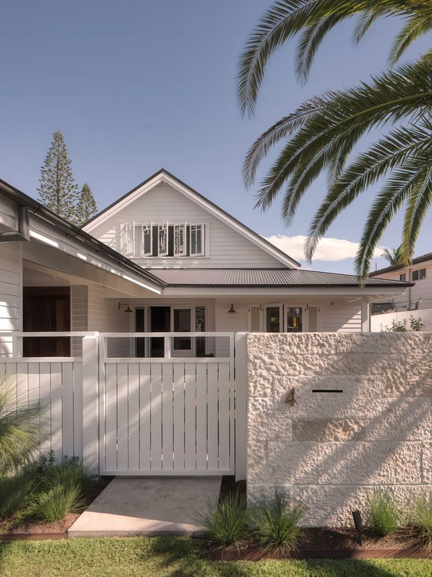 A bright exterior view of a white weatherboard house with a prominent pitched gable roof, corrugated metal roofing, upper-level windows, and a covered entry porch. A white slatted gate and textured rendered masonry boundary wall frame the front entrance, with palms and ornamental grasses in the landscaped foreground. The architecture has a relaxed coastal character and appears to be a multi-storey suburban residence.