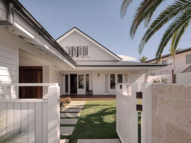 Front exterior view of a light-painted, single-storey home with a gabled roof, horizontal weatherboard cladding, covered entry and patio, white fencing, landscaped front path, and tall palm trees. The design has a relaxed coastal or traditional residential character.