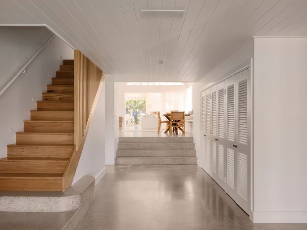 Bright, refined residential interior showing a polished concrete hallway and split-level transition. A warm timber staircase with vertical timber balustrade rises on the left, while full-height white louvred built-in storage lines the right wall. Steps lead up to a light-filled dining area with a timber dining table and chairs, and the interior features white-painted timber wall and ceiling panelling.