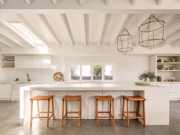 Bright, spacious white kitchen with a large central island and four timber bar stools. The room features white built-in cabinetry, a white subway-tile backsplash, open shelving, a broad multi-pane window, polished concrete flooring, and exposed white ceiling beams. Woven pendant lights add a warm natural accent.