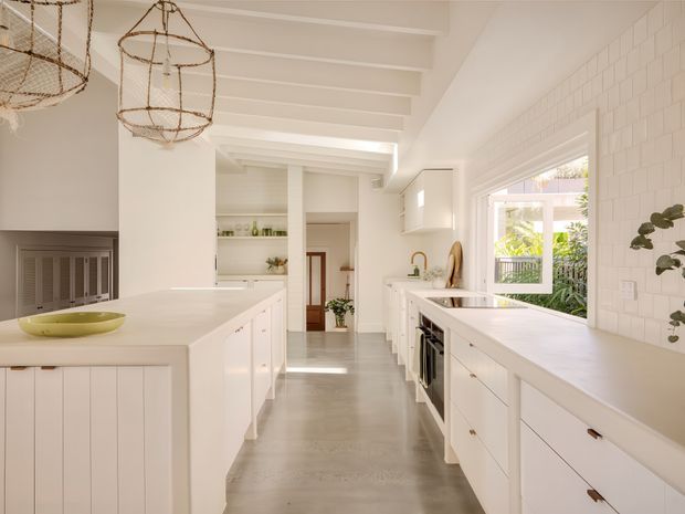Bright, spacious contemporary kitchen with white full-height cabinetry, two large islands, pale benchtops, a white tiled splashback, polished concrete flooring, exposed painted ceiling beams, and woven pendant lights. Large windows bring in abundant natural light and overlook greenery.