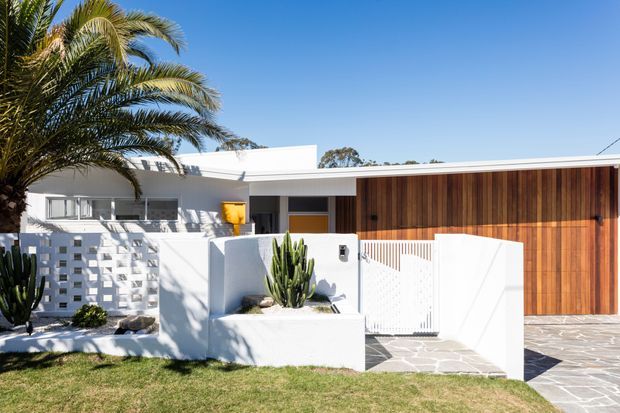 A bright, low-slung modern residence viewed from the street, featuring white rendered walls, a distinctive breeze-block privacy screen, warm vertical timber garage door cladding, a pale stone-paved driveway and entry, and sculptural drought-tolerant planting including palms and cacti.