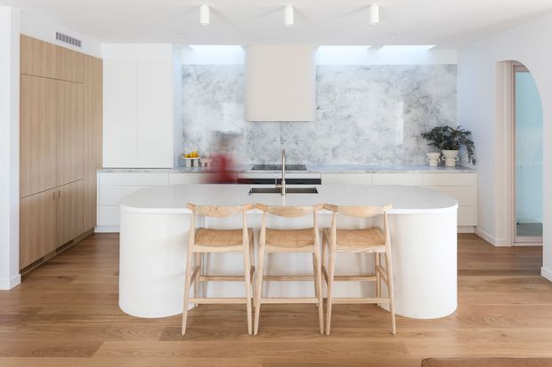 Bright contemporary kitchen featuring a large rounded white island with three timber bar stools, pale timber flooring, full-height light timber cabinetry, white cabinetry, and a veined grey-white stone backsplash. A concealed rangehood is integrated into the wall cabinetry, with a curved doorway leading to an adjacent blue-toned space.