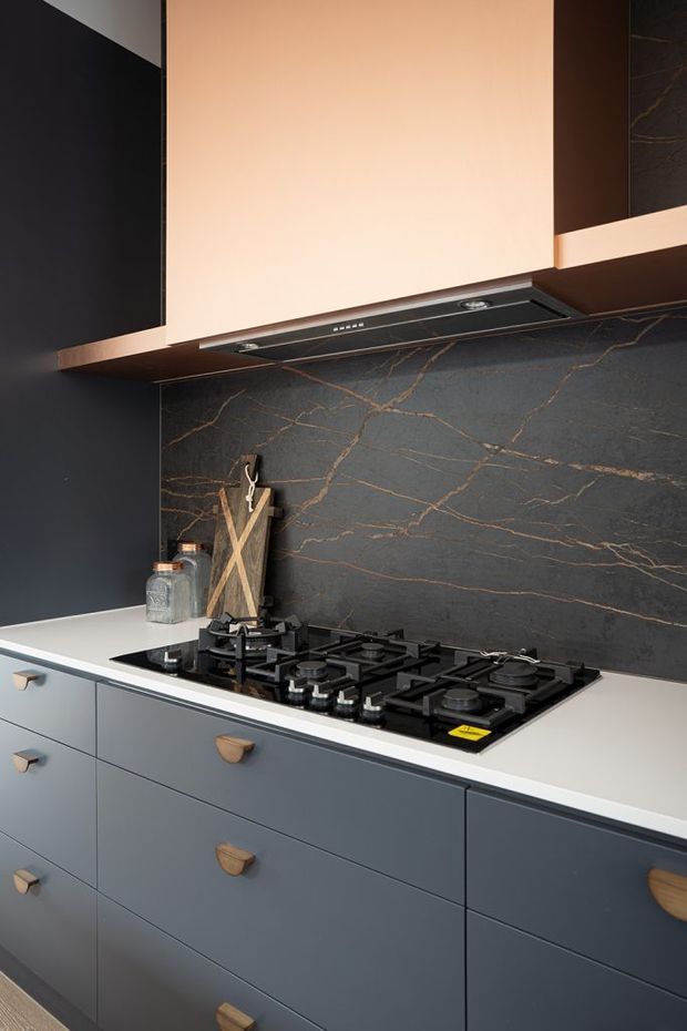 Close-up view of a contemporary kitchen cooking zone with dark blue-grey lower cabinetry, pale benchtop, integrated rangehood, black gas cooktop, and a dramatic dark stone-look splashback with fine bronze veining. Warm pale cabinetry above and timber-toned drawer pulls add contrast.