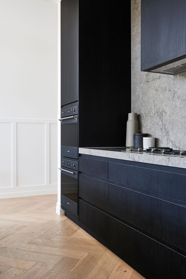 A close-up view of a refined contemporary kitchen featuring dark blue-black vertically textured cabinetry, integrated appliances, a pale grey veined stone benchtop and splashback, and a stainless steel cooktop. The kitchen opens to a bright adjoining space with white decorative wall panelling and warm herringbone timber flooring.