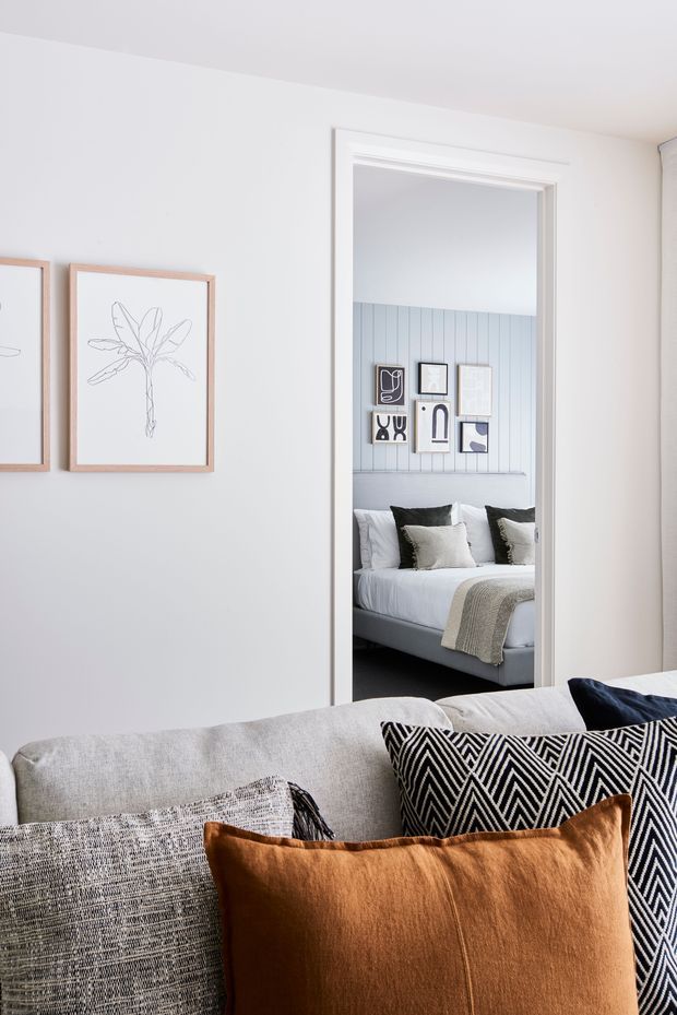 A bright, refined interior view from a living room toward an adjoining bedroom. The foreground features a light grey upholstered sofa with layered cushions in black-and-white geometric patterns, textured grey, and warm rust velvet. White walls display simple framed botanical artwork. Through the doorway, the bedroom has a pale blue vertical-panel feature wall, a neatly made bed with neutral bedding, and a curated gallery wall.
