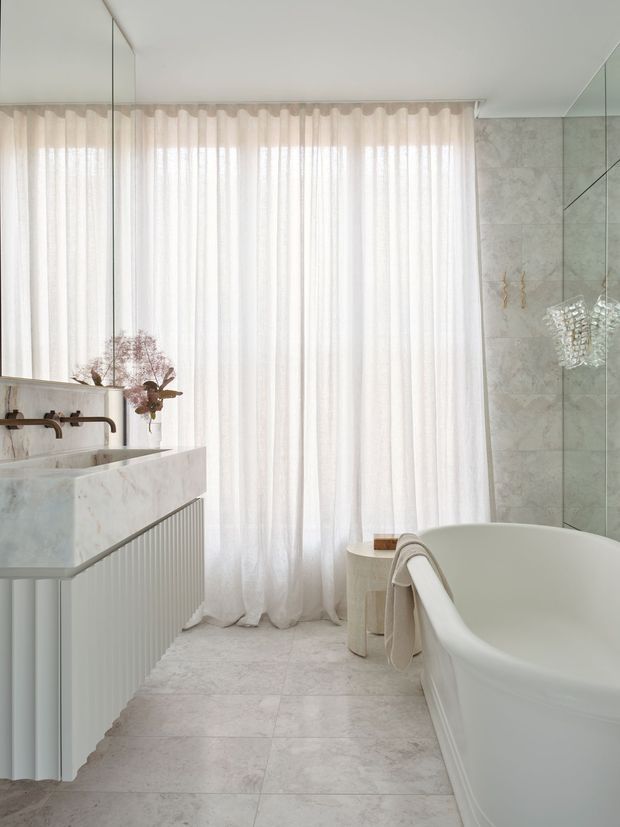 A bright, refined bathroom with a freestanding white bathtub, marble-look tiled walls and floor, a pale stone vanity with fluted cabinetry, brushed brass tapware, full-height sheer curtains, and mirrored surfaces. The soft white and pale grey palette creates an airy, calm atmosphere.