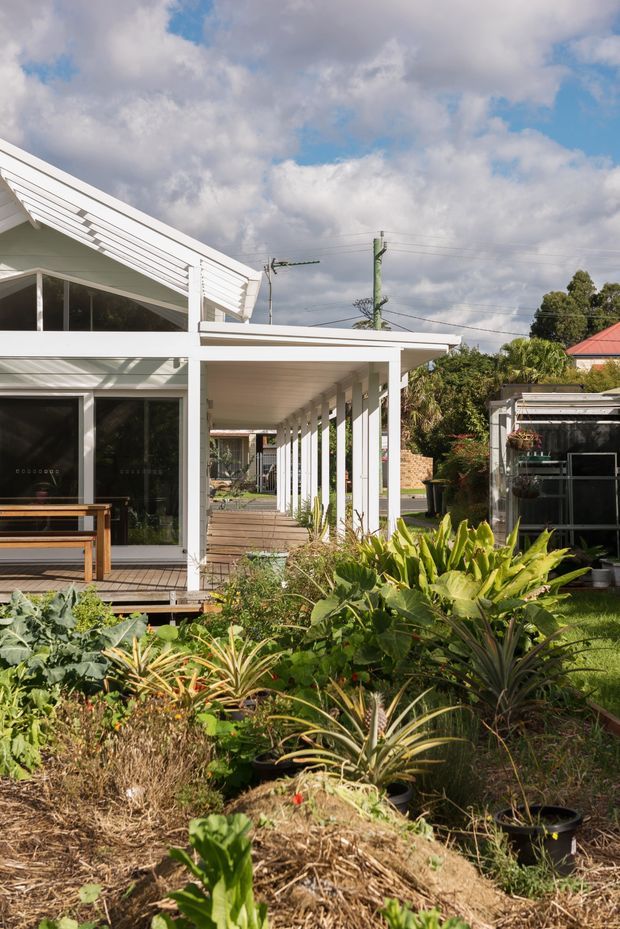 Exterior view of a low-rise white house with a prominent sloping roof, broad covered veranda, exposed white posts, and extensive glazing. A timber deck and outdoor bench sit beside the house, while a lush productive garden fills the foreground. The suburban setting includes neighbouring structures and utility lines beneath a partly cloudy sky.